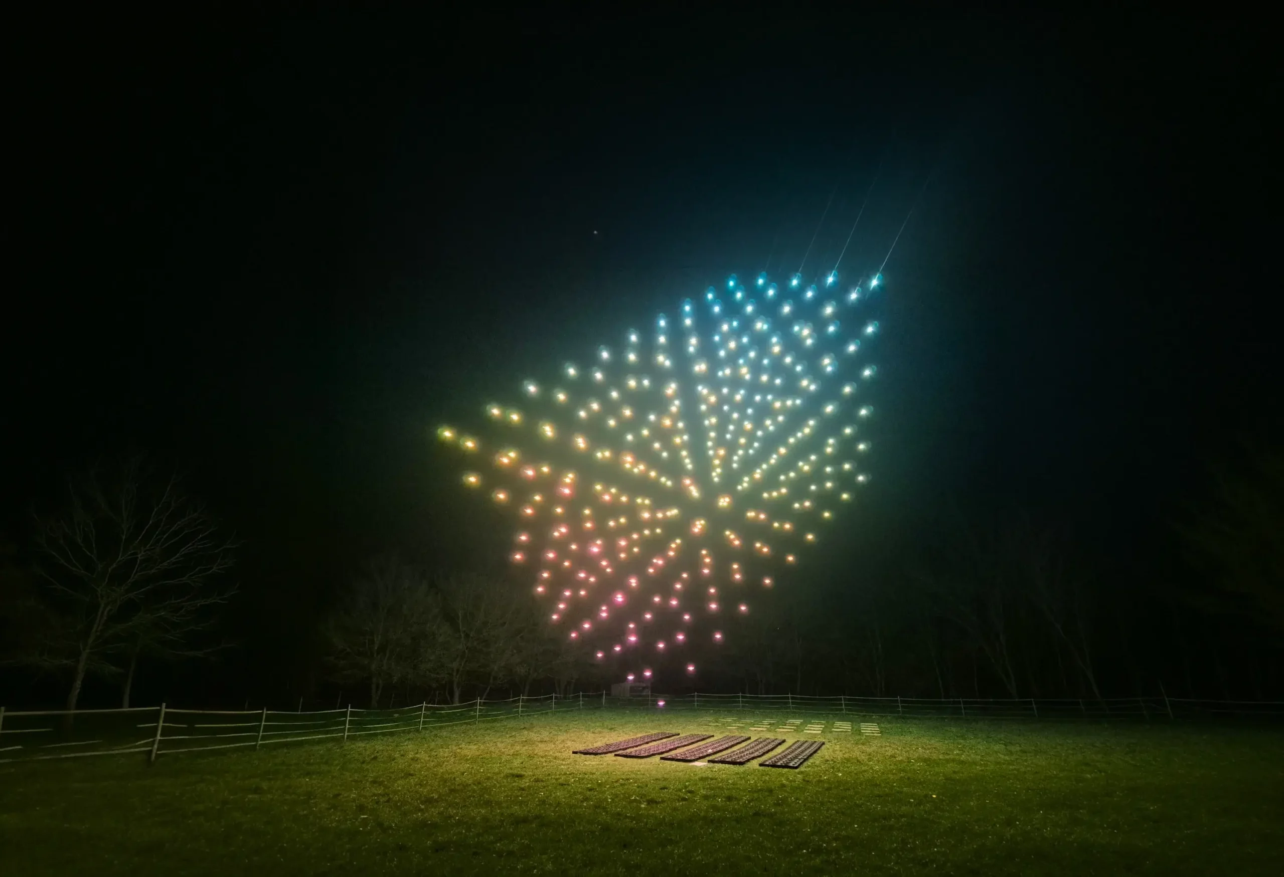 A synchronized swarm of illuminated drones forms a vibrant, multicolored geometric pattern in the night sky above a grassy field, with trees and ground equipment visible below.