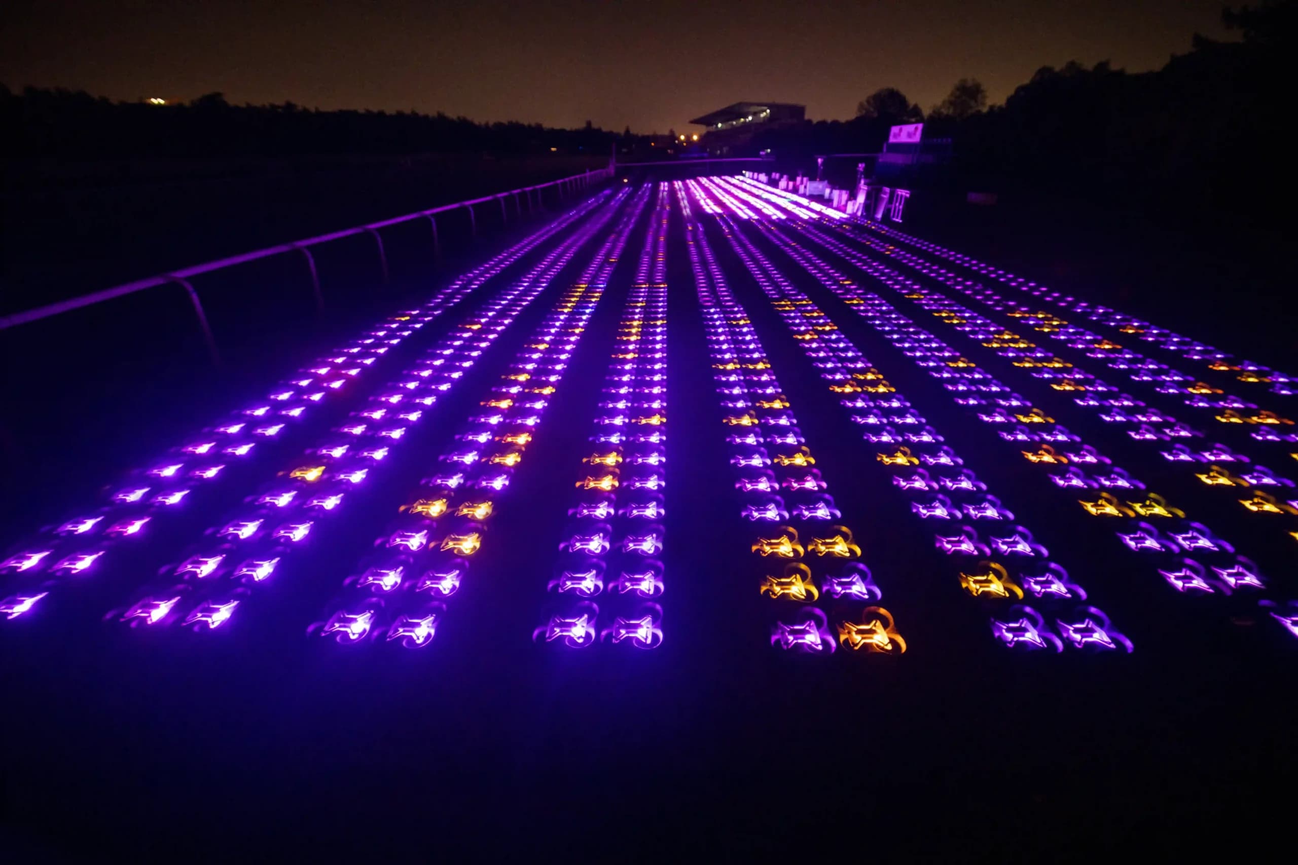 Hundreds of illuminated drones arranged in precise rows on the ground at night, glowing in purple and amber lights before a drone light show.