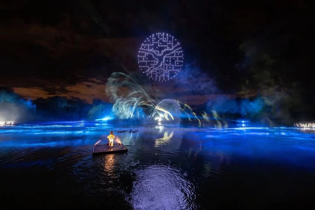 A nighttime drone light show over water, forming a glowing tree shape in the sky, with colorful light effects and reflections on the surface, while a performer stands on a small floating platform in the foreground.